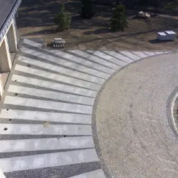 Widok z góry na nowo ułożony, półokrągły podjazd z szarej kostki betonowej przeplatanej ciemniejszą kostką granitową, prowadzący do domu.